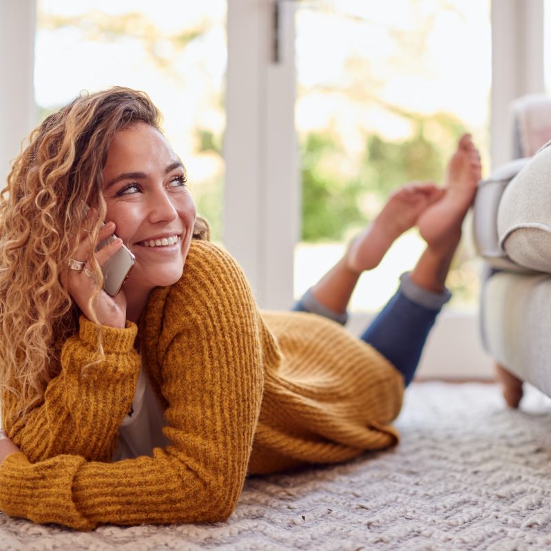 Woman In Warm Jumper Lying On Floor At Home Talking On Mobile Phone